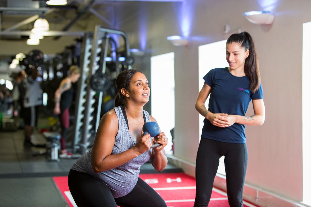 A lady in grey gym kit, using a kettlebell to do squats. A Personal Trainer is watching her.
